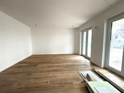 Das Bild zeigt eine leere Wohnecke mit hellem Holzboden und weißen Wänden. Zwei Türen und ein großes Fenster sorgen für viel Licht. An der Decke links sind freiliegende Kabel für Beleuchtung. Bodenleisten und Bauelemente liegen vorn rechts.