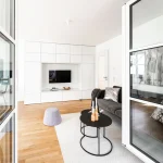 Helles, modernes Wohnzimmer mit Parkettboden; weiße Einbauschränke und TV Wand. Große Fensterfront und verglaste Schiebetür führen zu einem weiteren Raum. Graues Sofa, schwarzer Beistelltisch, grauer Pouf, weißer Teppich. Kunstwerk an der Wand.