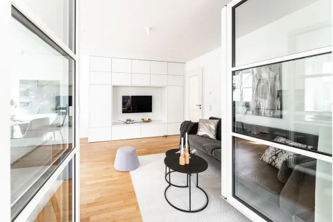 Modernes Wohnen: Stilvolles Zuhause mit klaren Linien Helles, modernes Wohnzimmer mit Parkettboden; weiße Einbauschränke und TV Wand. Große Fensterfront und verglaste Schiebetür führen zu einem weiteren Raum. Graues Sofa, schwarzer Beistelltisch, grauer Pouf, weißer Teppich. Kunstwerk an der Wand.