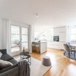 Ein geräumiges, helles Zimmer mit Wohn- und Essbereich. Links das Wohnzimmer mit grauem Sofa, schwarzem Couchtisch und Terrassenzugang. Rechts die weiße Küche mit Insel und die Essgruppe mit rundem Tisch und Stühlen. Helle Einrichtung und Holzfußboden.