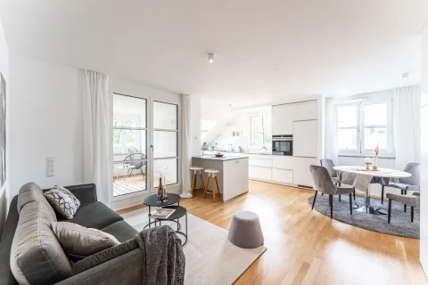 Helle Wohnung mit nahtlosem Wohnen-Essen Bereich Ein geräumiges, helles Zimmer mit Wohn- und Essbereich. Links das Wohnzimmer mit grauem Sofa, schwarzem Couchtisch und Terrassenzugang. Rechts die weiße Küche mit Insel und die Essgruppe mit rundem Tisch und Stühlen. Helle Einrichtung und Holzfußboden.
