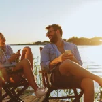Ein Mann und eine Frau sitzen entspannt auf Holzstühlen an einem See, möglicherweise Ammersee oder Starnberger See, genießen den Sonnenuntergang und schauen auf ihre Handys. Die Stimmung ist heiter und sie lachen zusammen.