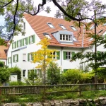 Mehrfamilienhäuser mit weißen Fassaden und grünen Fensterläden in Obermenzing. Umgeben von Bäumen und einem kleinen Garten. Ideal für Wohnungssuche in ruhiger, grüner Lage.