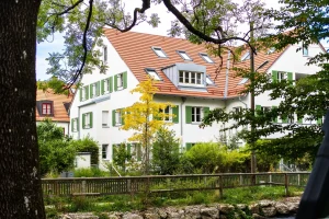 Idyllisches Wohnen in Obermenzing – Ihr neues Zuhause! Mehrfamilienhäuser mit weißen Fassaden und grünen Fensterläden in Obermenzing. Umgeben von Bäumen und einem kleinen Garten. Ideal für Wohnungssuche in ruhiger, grüner Lage.