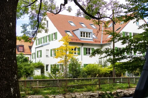Idyllisches Wohnen in Obermenzing – Ihr neues Zuhause! Mehrfamilienhäuser mit weißen Fassaden und grünen Fensterläden in Obermenzing. Umgeben von Bäumen und einem kleinen Garten. Ideal für Wohnungssuche in ruhiger, grüner Lage.