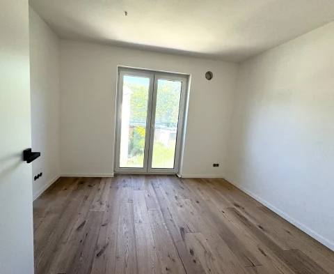Ein leerer Raum in einer Wohnung im Obergeschoss. Er hat weiße Wände, einen Holzfußboden und eine Terrassentür, durch die man auf einen grünen Garten blickt. Der Raum ist hell erleuchtet und unmböbliert.