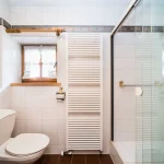 Das Bild zeigt ein modernes, helles Badezimmer. Es enthält eine Toilette, ein Bidet, einen Heizkörper und eine große Duschkabine mit Glaswänden. Über dem Bidet und der Toilette befindet sich ein Fenster mit weißer Gardine.